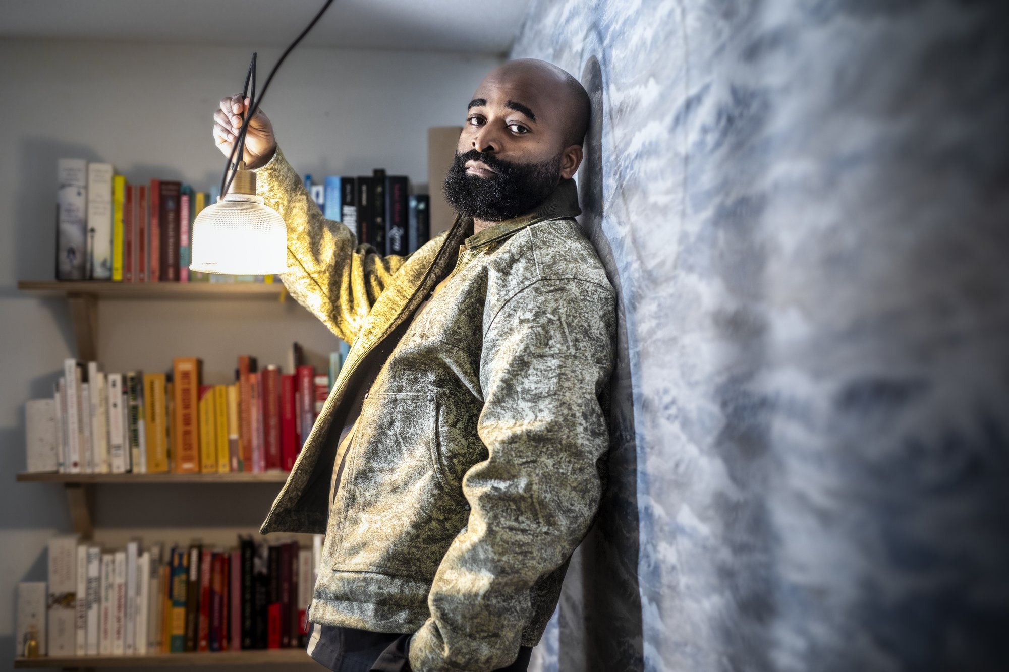 Kiza Magendane bij een lamp, leunend tegen een muur naast een boekenkast