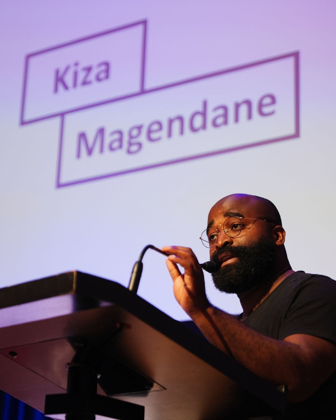 Kiza Magendane bij het katheder, met zijn naam op het scherm achter hem